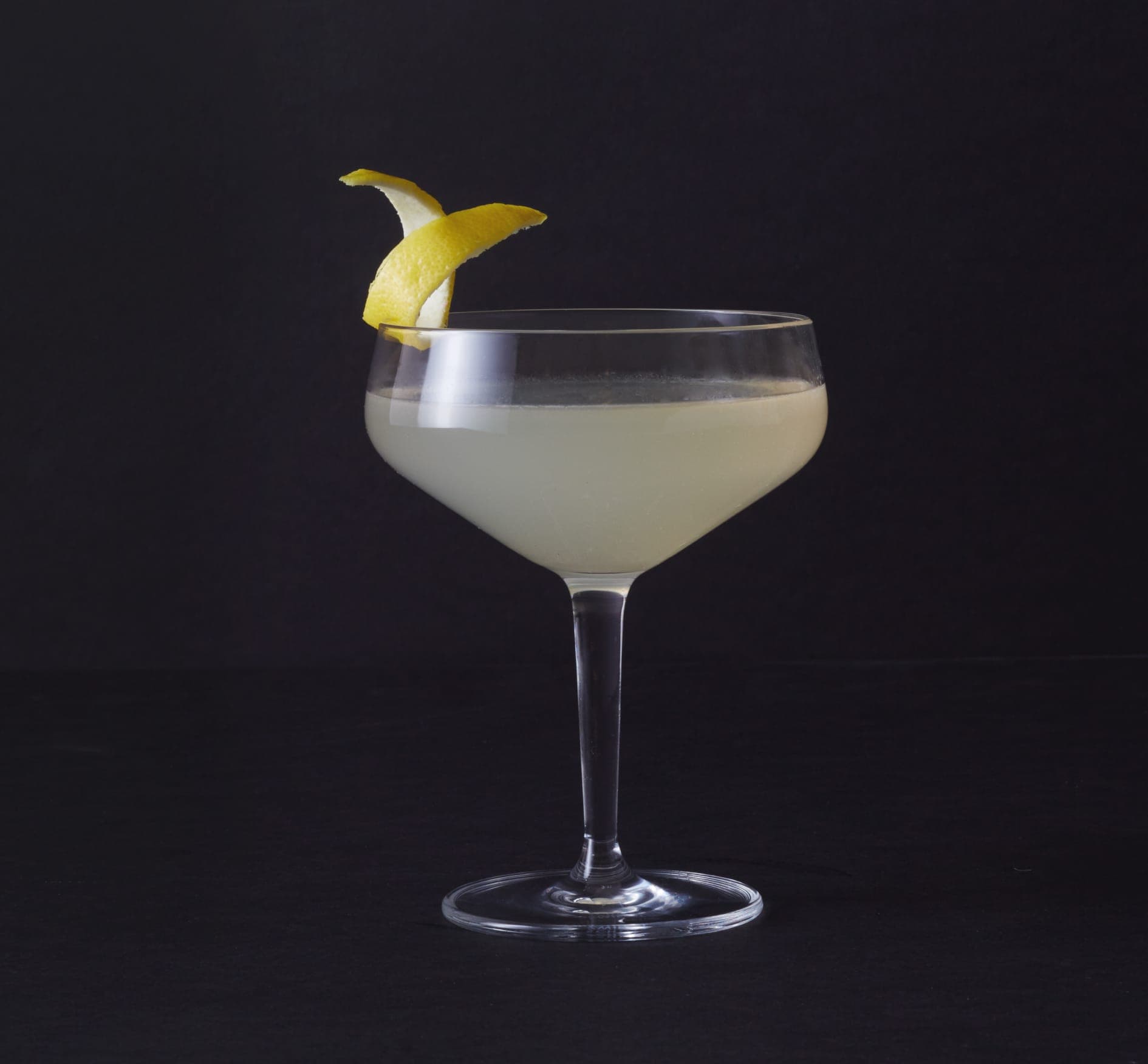 A cocktail glass sits in the centre on a black background. The glass is filled with a liquid the colour of lemon juice and is slightly cloudy. The rim of the glass is garnished with a twist of lemon peel.